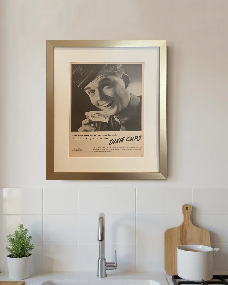 Original Dixie Cups vintage advertisement beautifully framed in brushed brass with an off-white mat, displayed in a bright modern kitchen.
