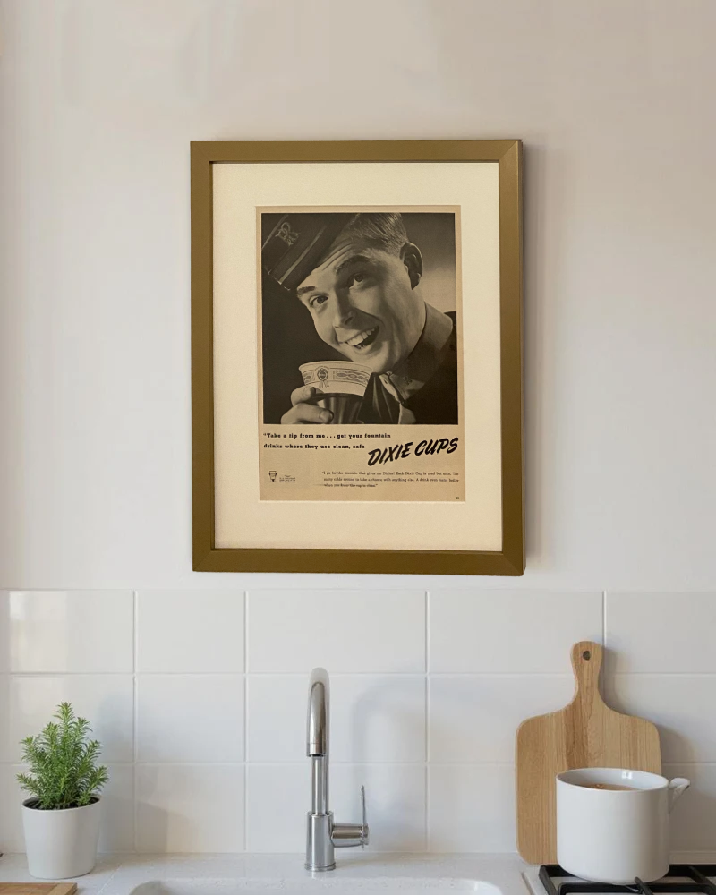 Original vintage Dixie Cups advertisement framed in medium brass and styled on a modern kitchen wall above a white tile backsplash.