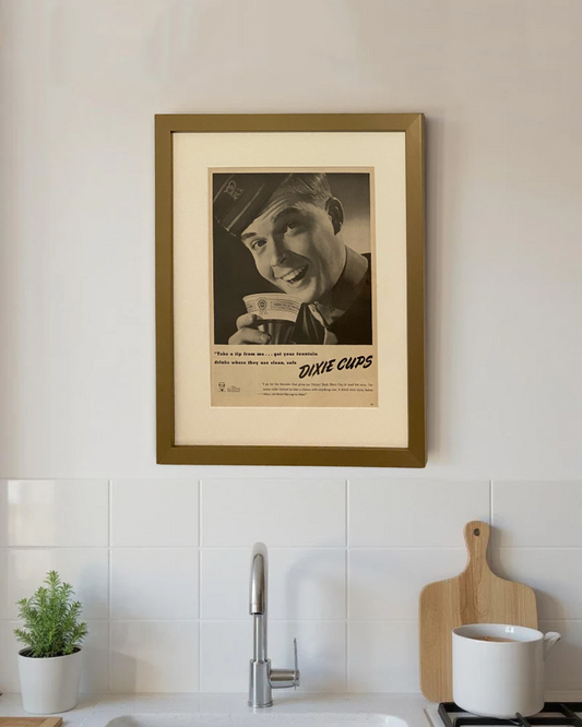 Original vintage Dixie Cups advertisement framed in medium brass and styled on a modern kitchen wall above a white tile backsplash.