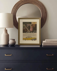Original 1955 Cadillac vintage advertisement displayed in a thin brass frame on a bedroom dresser with warm modern décor.