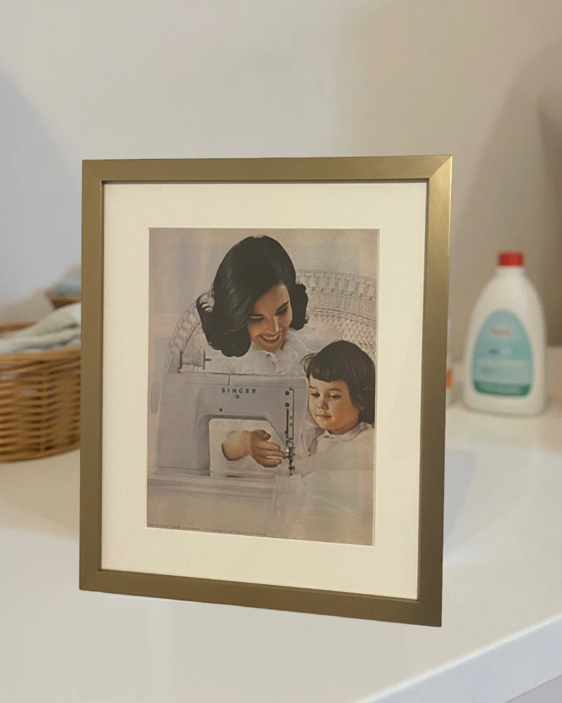 Original 1960s Singer “Mother and Daughter” magazine advertisement professionally framed in a matte brass frame on a modern bedroom nightstand.