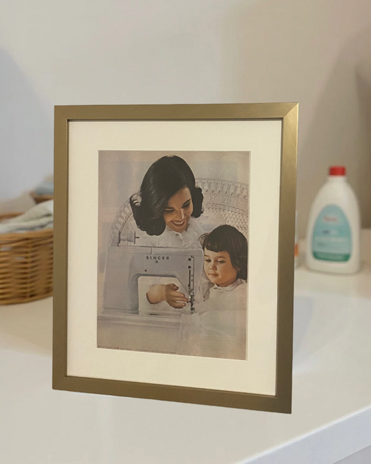Original 1960s Singer “Mother and Daughter” magazine advertisement professionally framed in a matte brass frame on a modern bedroom nightstand.